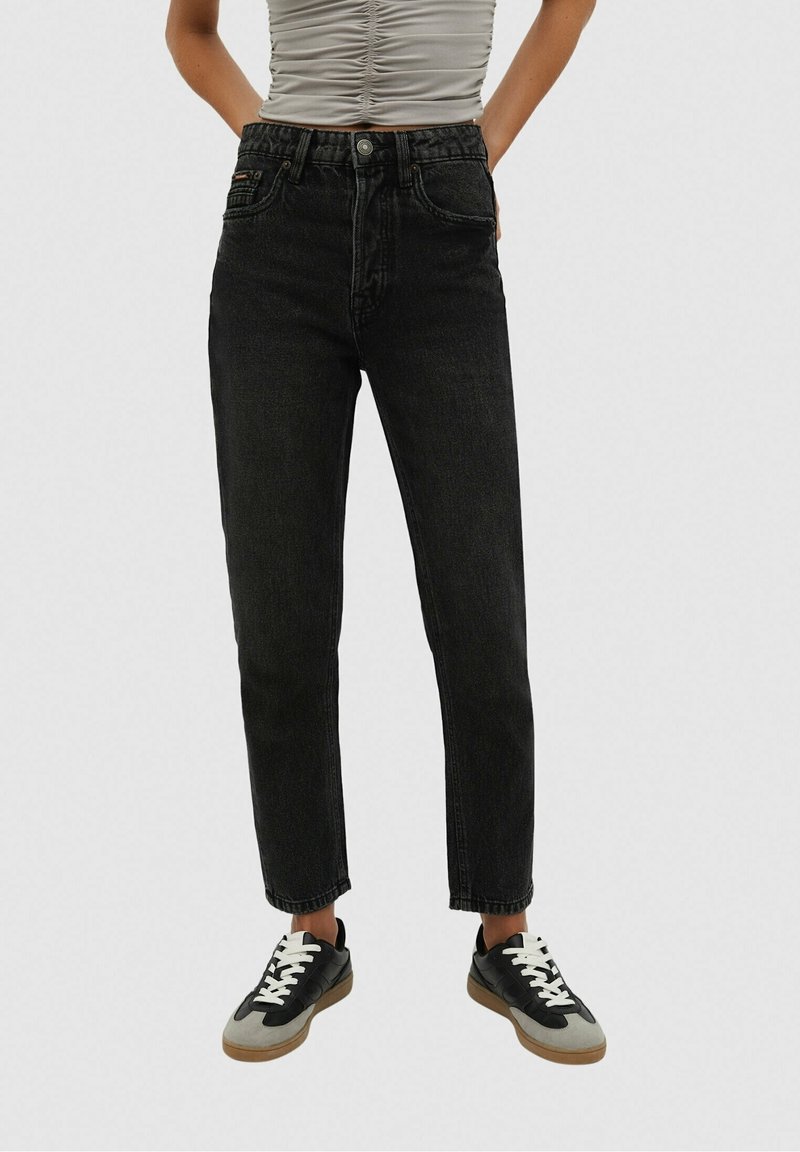 Personne portant un jean noir taille haute, un haut sans manches léger froncé et des baskets noires et grises à lacets blancs, debout les mains dans les poches.