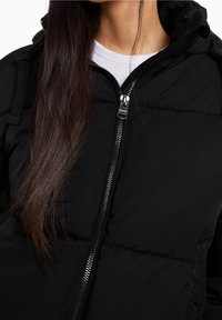 Černá polstrovaná bunda s vysokým límcem, zipovým zapínáním a texturovanou látkou. Obsahuje zip a nastavitelné popruhy na ramenech.
