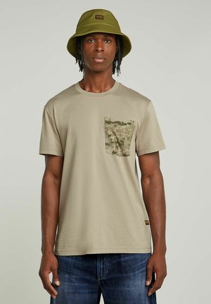 Beige katoenen T-shirt met een getextureerde camouflagezak, ronde halslijn en korte mouwen, gecombineerd met blauwe denim jeans en een groene bucket hat.