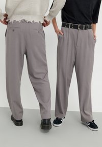 Gray tailored trousers with a relaxed fit, featuring double back pockets, and worn with a black belt. Two pairs of shoes visible.