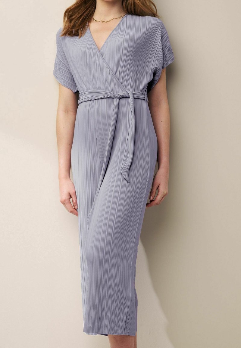 Vestido cruzado plisado en lavanda claro con mangas cortas, escote en V y cinturón atado a la cintura. Cae por debajo de la rodilla y presenta una textura vertical.