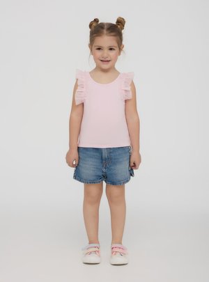 Młoda dziewczyna stoi, ubrana w różowy top bez rękawów z falbankami na ramionach, dżinsowe szorty i różowe sneakersy z podwójnymi paskami.