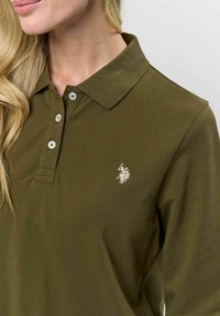 Olíva zöld pamut póló, lapos gallérral, három gombbal, és egy kis fehér hímzett logóval a bal mellrészen.