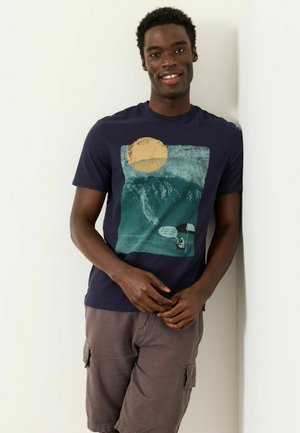 Young man smiling, leaning against a wall, wearing a dark blue graphic t-shirt with a sun and mountain design and brown cargo shorts.