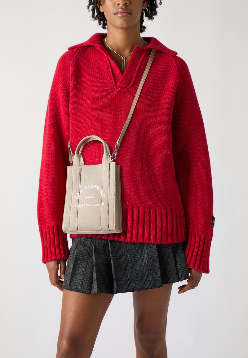 Rød strikket genser med bred krage, parret med en liten beige toteveske med brodert tekst. Pliert svart rutet skjørt fullfører antrekket.