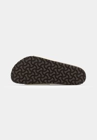 Semelle d'une sandale avec un motif en caoutchouc noir texturé comportant des formes courbes entrelacées sur une base beige.