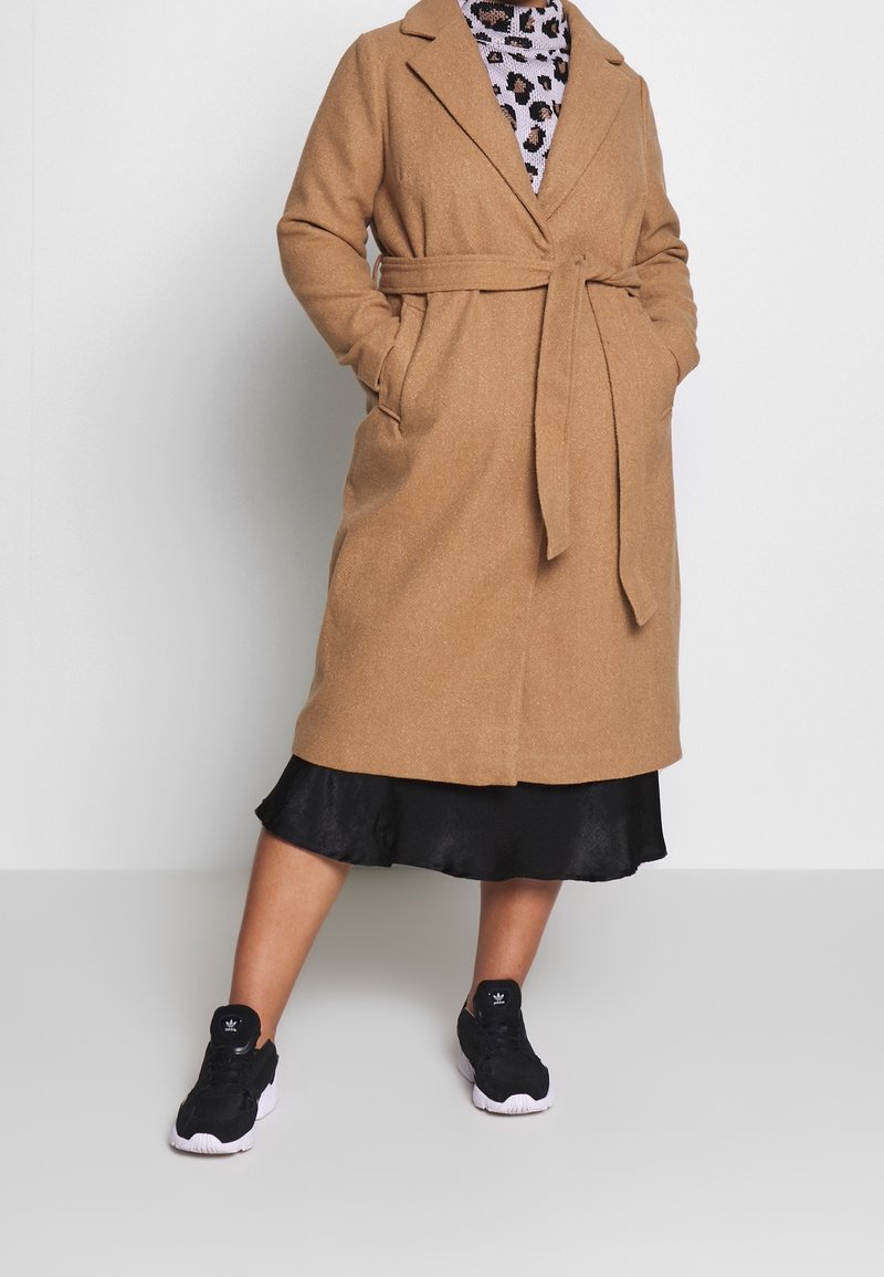 Long manteau marron en mélange de laine avec une ceinture, des poches latérales et un col cranté. Porté sur une robe noire et des baskets noires.
