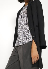 Czarna, dopasowana marynarka na dwa guziki, noszona na wzorzystej bluzce w jasnych odcieniach z ciemnymi geometrycznymi kształtami, zestawiona z szarymi spodniami.