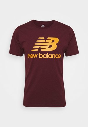 T-shirt en coton marron avec manches courtes, présentant un grand logo "NB" jaune et les mots "new balance" imprimés en dessous en jaune.