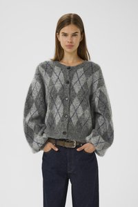 Woman wearing a gray fuzzy cardigan with diamond patterns and black buttons, paired with dark jeans and a brown belt, hands in pockets.