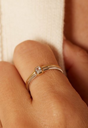 Gold and silver dual-band ring with a square clear gemstone worn on a person's finger against a cream fabric background.