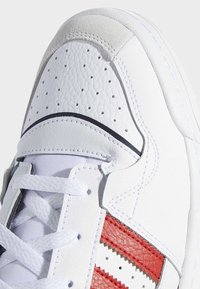 Gros plan sur l'avant d'une basket en cuir blanc avec perforations, lacets blancs et bandes rouges texturées sur le côté.