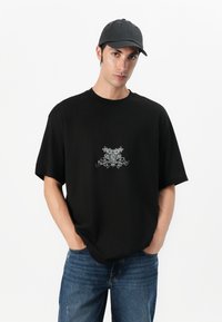 Black cotton t-shirt with short sleeves and a loose fit, featuring a silver graphic design on the chest, paired with blue denim jeans.