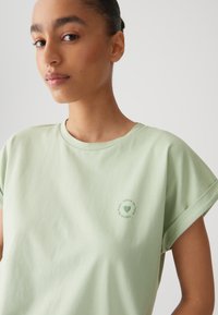 T-shirt in cotone verde chiaro con maniche corte arrotolate. Presenta una grafica circolare con testo verde vicino alla spalla sinistra. Texture morbida.