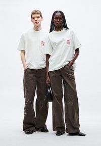 Dos modelos con camisetas de gran tamaño color gris claro con gráficos rojos y bolsillos tipo parche, combinadas con pantalones anchos marrones y zapatos negros.