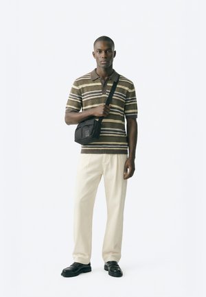 Moški, oblečen v rjavo polo majico s kratkimi rokavi in črtami, krem hlače, črne usnjene čevlje ter črno torbico čez ramo na enobarvnem belem ozadju.