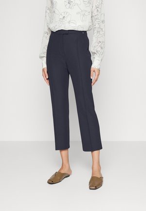 Pantaloni cropped blu navy con una vestibilità sartoriale, caratterizzati da una texture liscia e una parte anteriore piatta. Abbinati a una camicetta a fantasia e scarpe slide marroni.
