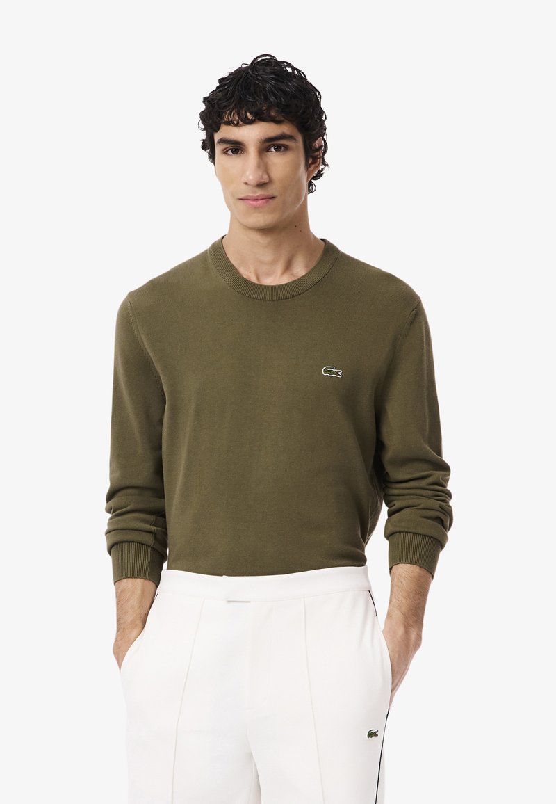 Joven vistiendo una camisa de manga larga verde oliva y pantalones blancos, de pie con las manos en los bolsillos frente a un fondo liso.