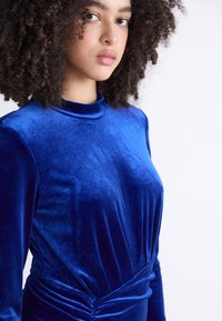 Vestido longo de veludo azul com mangas longas e gola alta, apresentando tecido franzido na cintura e uma textura suave e macia.