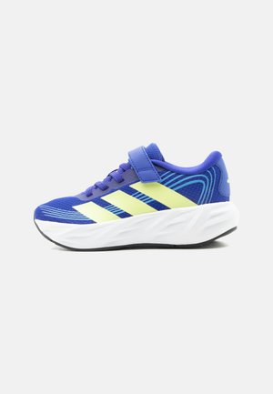 Chaussure de sport bleue avec des rayures vert clair, lacets et bride à velcro, semelle blanche rembourrée et semelle extérieure noire.