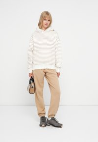 Hanorac crem cu dungi bej, croială lejeră. Asortat cu pantaloni de trening bej și adidași gri. Ținând o geantă mică neagră.