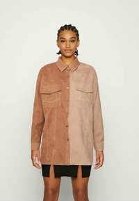 Camisa de botão em dois tons, castanho e bege, feita de material suave e texturizado, com dois bolsos no peito e detalhes de costura em contraste.