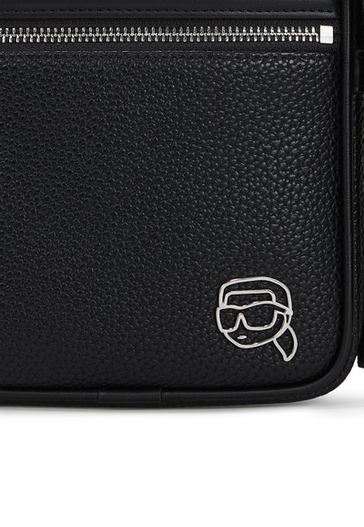 Sac en cuir noir texturé avec une fermeture éclair en métal argenté. Il présente un design minimaliste en relief d'un visage portant des lunettes de soleil sur le devant.