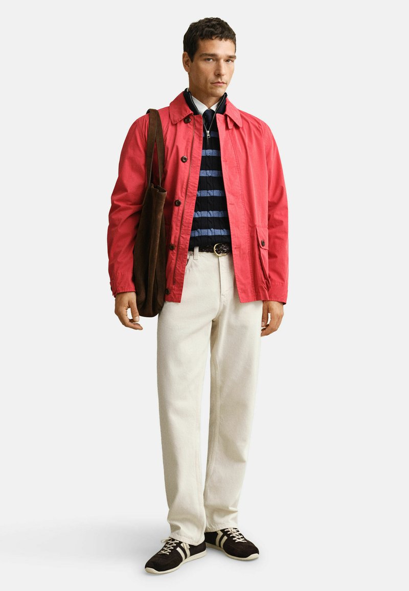 Homme portant une veste rouge, un pull à rayures bleu marine, un pantalon blanc, des baskets noires, et portant un sac à bandoulière marron, debout devant un fond blanc.