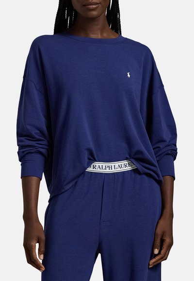 Γυναίκα που φορά μπλε μακρυμάνικη μπλούζα και παντελόνι στο ίδιο χρώμα, με ορατή λευκή ελαστική ζώνη μέσης με την ένδειξη "Ralph Lauren".