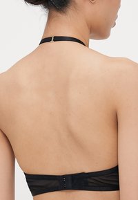 Zwarte halter bralette, van doorschijnend gaasmateriaal, verstelbare rugriem met gouden hardware, eenvoudige haaksluiting en strakke lijnen.
