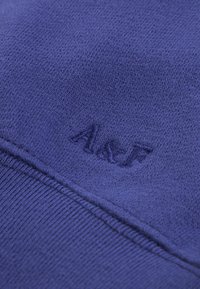 Primer plano de un tejido de punto púrpura con el acrónimo bordado en azul marino "AAIF" y una costura visible debajo del bordado.