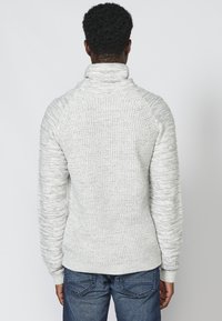 Hellgrauer Pullover mit hohem Kragen, strukturiertem Strickmuster und gerippten Bündchen, kombiniert mit blauen Jeans. Rückansicht.