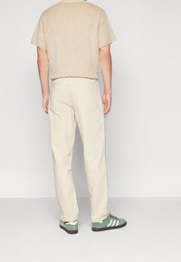 Camisa de punto texturizada beige, pantalones crema y zapatillas verdes con rayas blancas. Vista trasera, con dos bolsillos traseros en los pantalones.