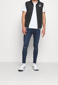Chaleco de plumas negro sobre una camiseta blanca, combinado con jeans ajustados de color azul oscuro con roturas en las rodillas, y zapatillas deportivas en negro y blanco.