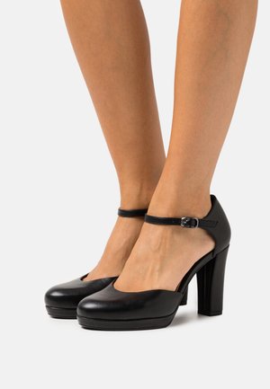 Černé kožené boty na vysokém podpatku s kulatou špičkou, páskem kolem kotníku a přezkou. Mají platformu pro zvýšení výšky a stability.