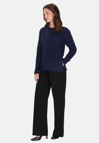 Granatowy sweter w warkoczowy wzór z okrągłym dekoltem, długimi rękawami i bocznymi rozcięciami, połączony z czarnymi spodniami o szerokich nogawkach i czarnymi butami.