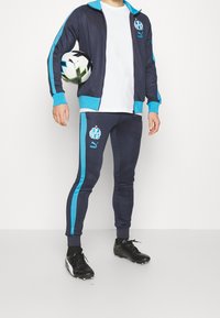 Námořnický tréninkový komplet s tyrkysovými akcenty, zahrnující zipovou bundu a těsné kalhoty. Černé fotbalové kopačky. Model držící fotbal.