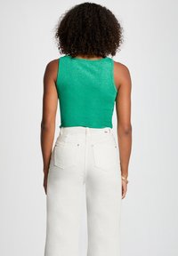 Groene, mouwloze top met een glinsterende textuur en een ronde halslijn, gecombineerd met hoge witte jeans met achterzakken en knoopaccenten.