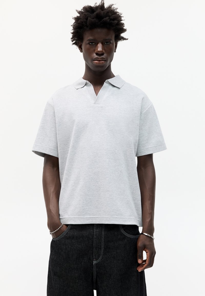 Grå polo t-shirt med krave og korte ærmer. Lavet af blødt stof, har en v-hals åbning og en afslappet pasform. Bæres med mørke denimbukser.