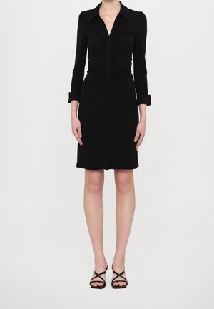 Robe noire à manches longues avec col, détails froncés, boutonnage avant et ourlet jusqu'aux genoux. Portée avec des sandales noires à lanières.