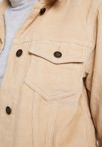 Veste en velours côtelé beige avec un tissu texturé, dotée d'un col pointu, de deux poches poitrine et d'accents en boutons noirs.
