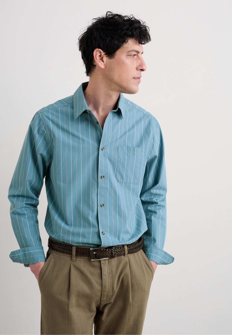 Lichtblau gestreepte shirt met een kraag, knoopsluiting aan de voorkant en een zak op de linkerborst. Gecombineerd met beige geplooide broeken en een geweven bruine riem.