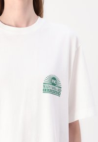White cotton t-shirt with a round neckline and short sleeves, featuring a green logo print on the left chest that includes text and sunburst design.