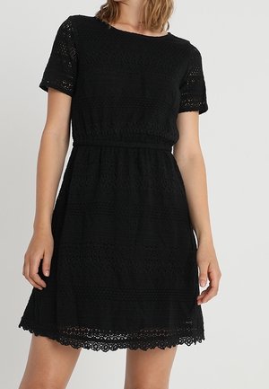Kobieta w krótkiej czarnej koronkowej sukience z wcięciem w talii i falowanym dołem na jednolitym tle.