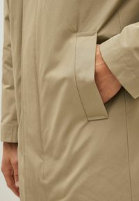 Beige vandtæt frakke med en glat tekstur, der har en sidelomme og synlige syninger langs sømmene. Modelens hånd hviler i lommen.