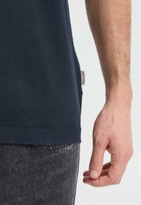 Pull en tricot marine avec ourlet côtelé, étiquette latérale, porté avec un jean gris. Gros plan sur une main reposant contre le côté du pull.