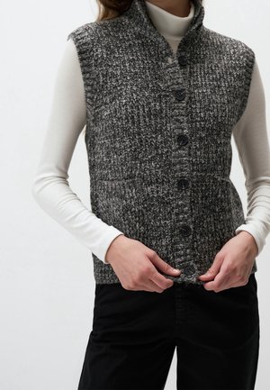 Femme portant un gilet en maille grise texturée à boutons par-dessus un col roulé blanc à manches longues et un pantalon noir, ajustant le bouton inférieur du gilet.