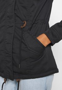 Chaqueta de tela negra con cintura ajustable, puños elásticos, bolsillos frontales y detalles en marrón en los toggles y los extremos del cordón.