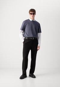 Pilkos spalvos apykaklė t-shirt, uždėta ant balto dryžuoto ilgomis rankovėmis marškinio, juodos kelnės ir juodi batai. Modelis dėvi tamsius akinius nuo saulės.