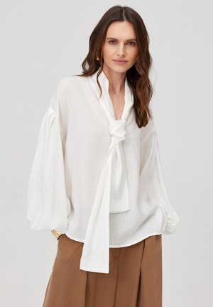 Kobieta z długimi falowanymi brązowymi włosami, nosząca złote kolczyki koła, białą bluzkę z przesadnie szerokimi rękawami i wiązaniem z przodu oraz brązowe spodnie z wysokim stanem.